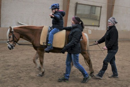Foto: Ein Schüler mit körperlicher Beeinträchtigung sitzt auf einem Pferd. Das Pferd wird von hinten am Langzügel geführt und geht im Schritt. Eine Therapeutin begleitet die beiden.