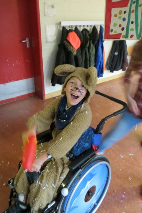 Auf dem Bild sieht man einen als Hase verkleideten Schüler fröhlich mit dem Rollstuhl im Karnevalszug durch die Schule fahren.