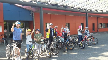Schülerinnen und Schüler stehen mit ihren Fahrrädern, Dreirädern und Liegerädern mit einer Polizistin und einem Polizisten strahlend vor der Schule und machen "Daumen hoch" Zeichen.