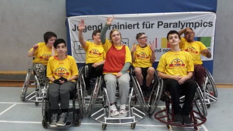 Ein Gruppenfoto in gelben Trikots in den Sportrollstühlen vor einem Plakat mit der Aufschrift "Jugend trainiert für Olympia".