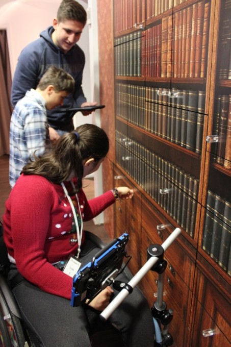 Foto: Eine Schülerin im Rollstuhl, ein Schüler und ein Erwachsener stehen mit iPad vor der Bücherregal mit einem Tresor.